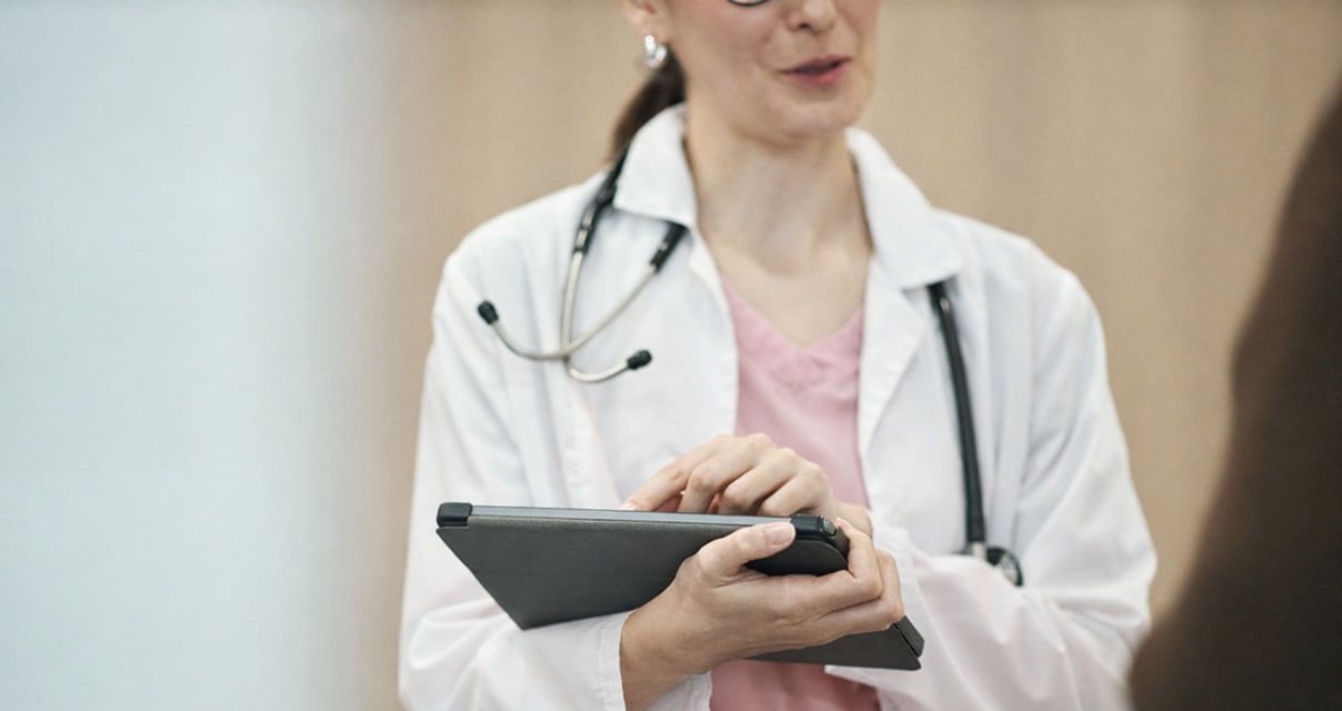 A doctor wearing a white coat and stethoscope holds a tablet and speaks with someone off-camera, appearing to review or explain information.