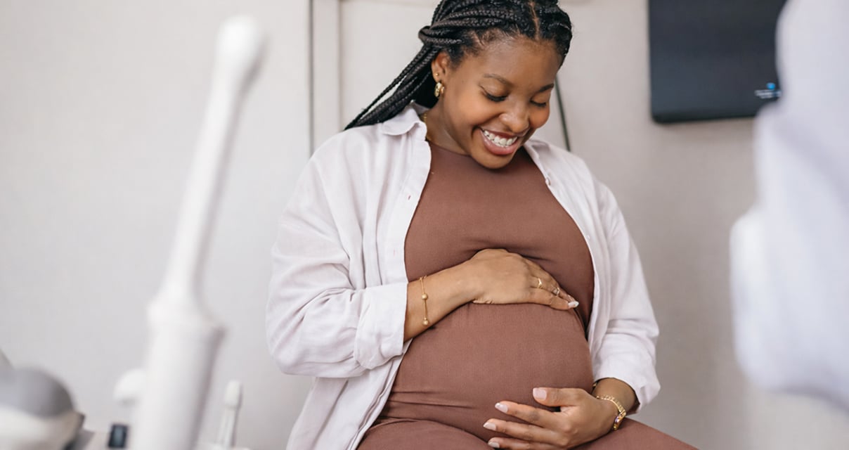 A pregnant woman smiling and cradling her belly with both hands while sitting in a medical clinic, wearing a brown dress and white shirt.