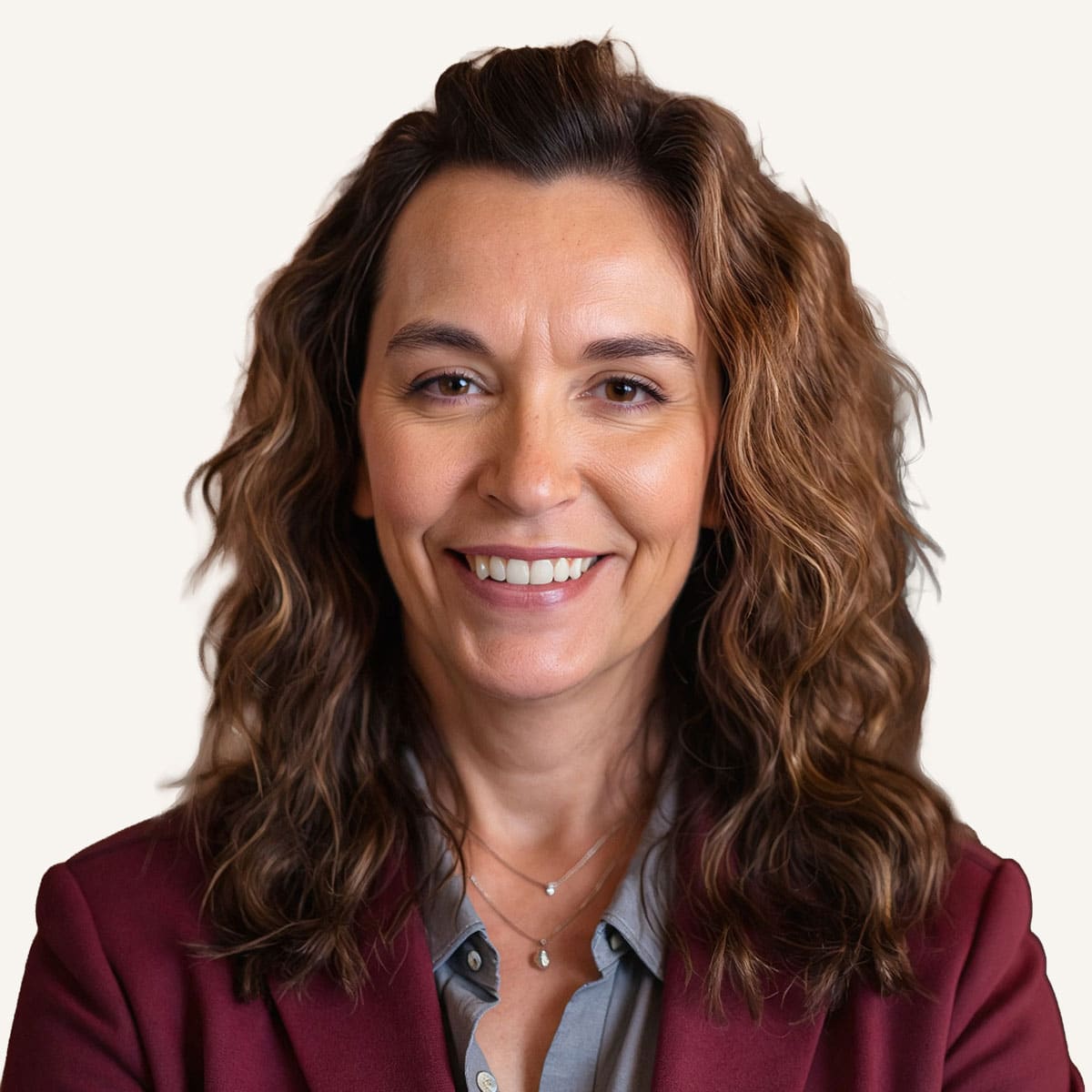 A woman with wavy brown hair wearing a maroon blazer, gray shirt, and a silver necklace, smiling at the camera against a plain light background.