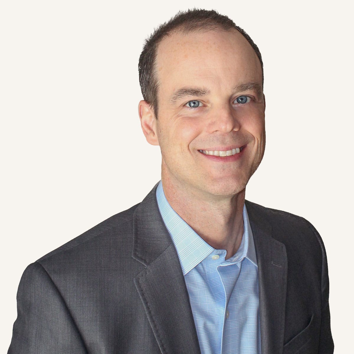 A man with short brown hair, wearing a gray suit jacket over a light blue collared shirt, smiles while looking at the camera against a plain light background.
