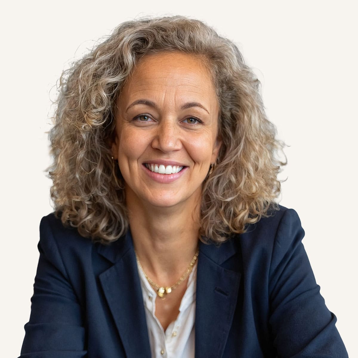 A smiling woman with curly gray hair wearing a dark blazer and white blouse, sitting and facing the camera against a light background.