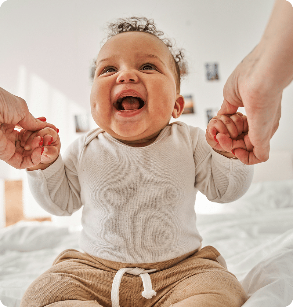 A smiling baby sits on a bed, wearing a white long-sleeve shirt and beige pants, holding onto two adult hands for support. The room is bright with sunlight and some photos on the wall in the background.