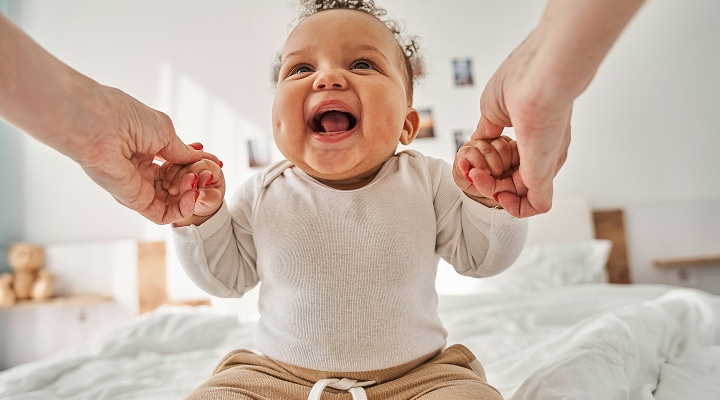 Smiling baby sitting on a bed, holding the hands of an adult on each side. The baby looks happy and excited, wearing a light-colored long-sleeve shirt and brown pants in a bright, cozy bedroom.