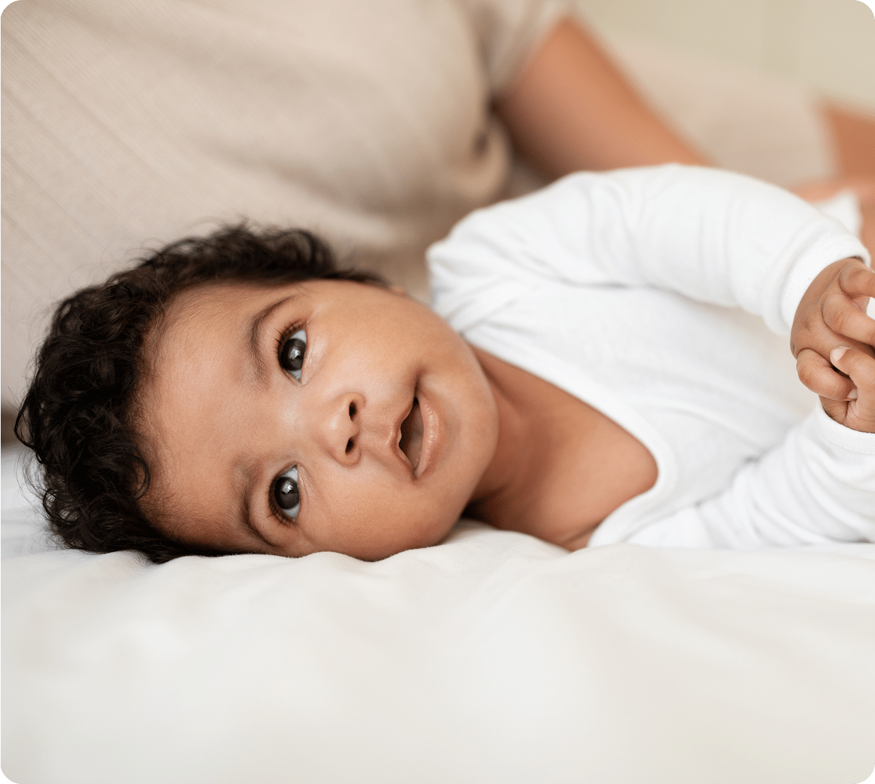 A baby with curly dark hair lies on a white bed, wearing a white onesie. The baby looks up with wide eyes and a gentle expression, with an adults arm visible in the background.