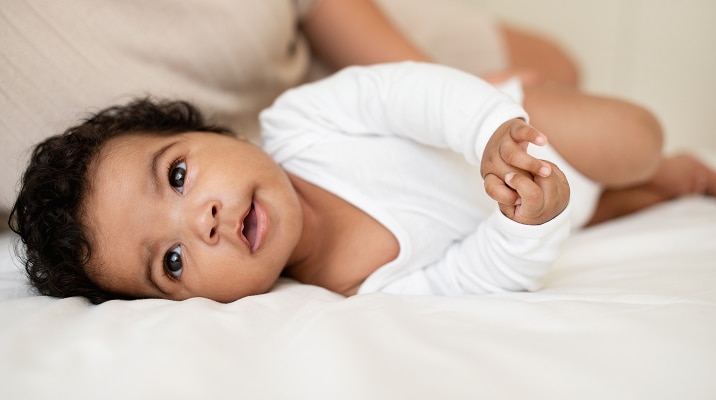 A baby in a white onesie lies on a bed, looking up with a curious expression. An adults arm is partially visible in the background, gently supporting the baby.