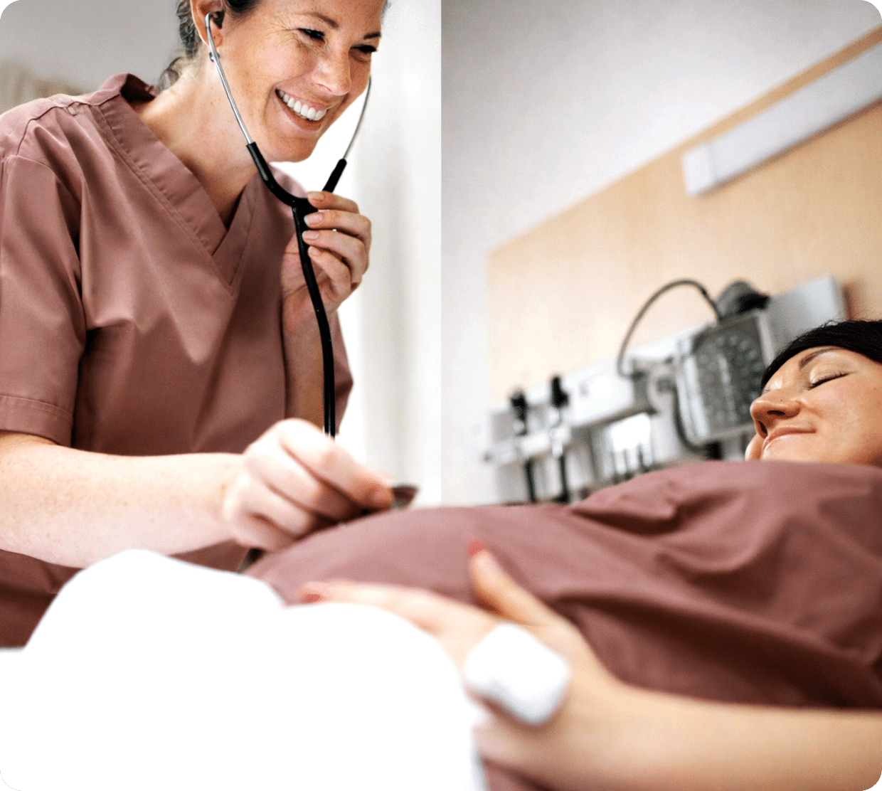 A smiling healthcare worker in scrubs uses a stethoscope to check a pregnant womans belly as she lies on a hospital bed, both appearing relaxed in a clinical setting.
