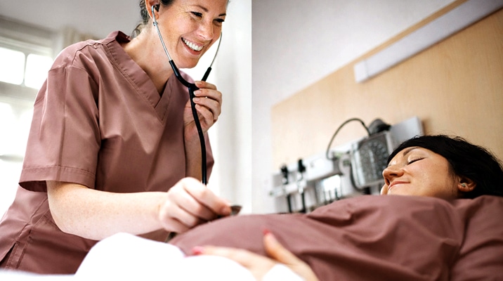 A smiling healthcare worker uses a stethoscope to check a pregnant woman lying in a hospital bed. The patient is relaxed, and medical equipment is visible on the wall behind her.