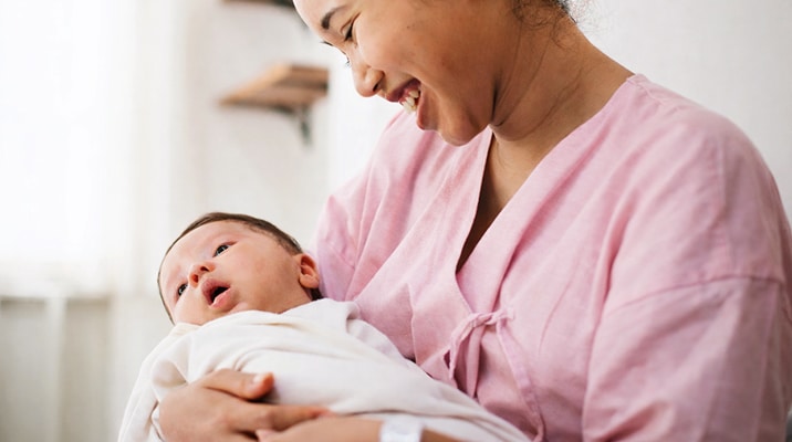 A woman in a pink top smiles warmly while holding a newborn baby wrapped in a white blanket. The baby looks up, and natural light fills the room, creating a gentle, peaceful atmosphere.