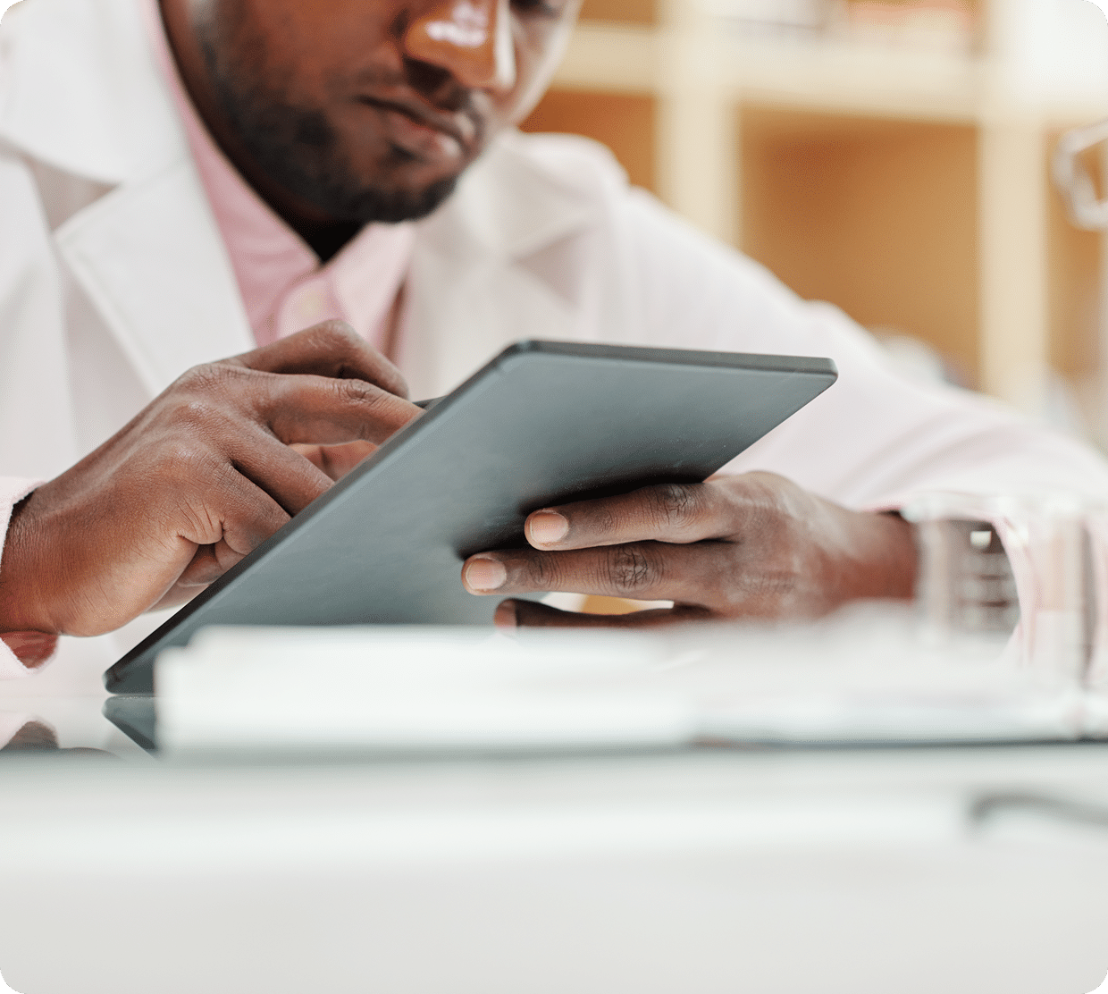 A person wearing a white lab coat uses a tablet device at a desk, with shelves and blurred objects in the background. Only their hands and part of their face are visible.