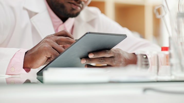 A person in a white lab coat uses a tablet device, with laboratory equipment blurred in the foreground.